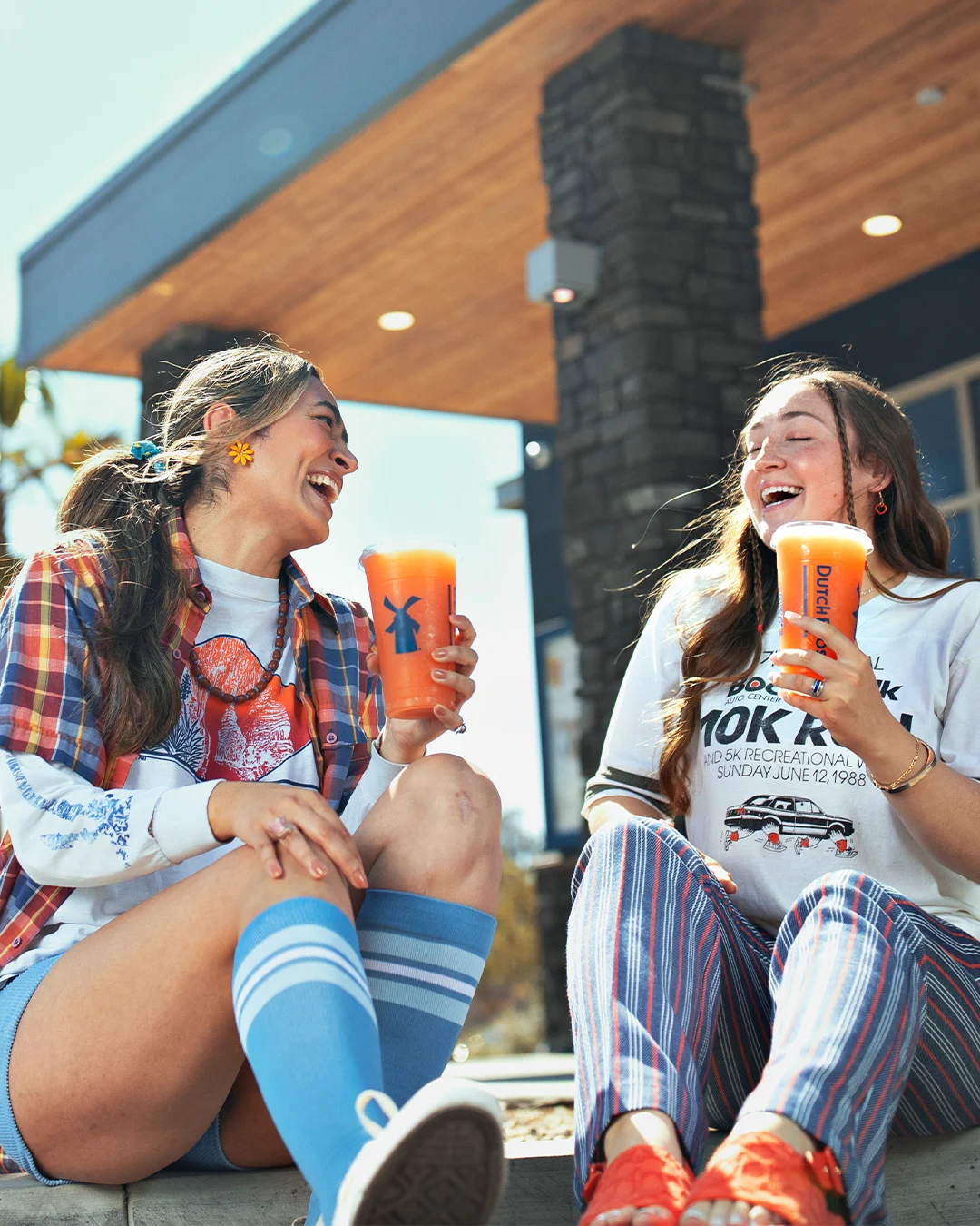 Two friends laughing while enjoying orange drinks from Dutch Bros outside a café with wooden overhang.