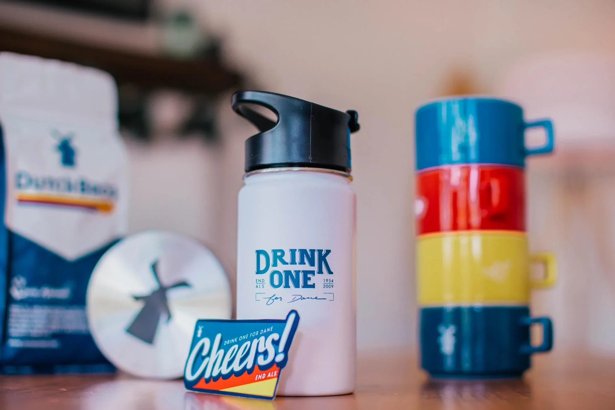 White water bottle with "DRINK ONE" text next to colorful travel mug and Dutch Bros coffee packaging.