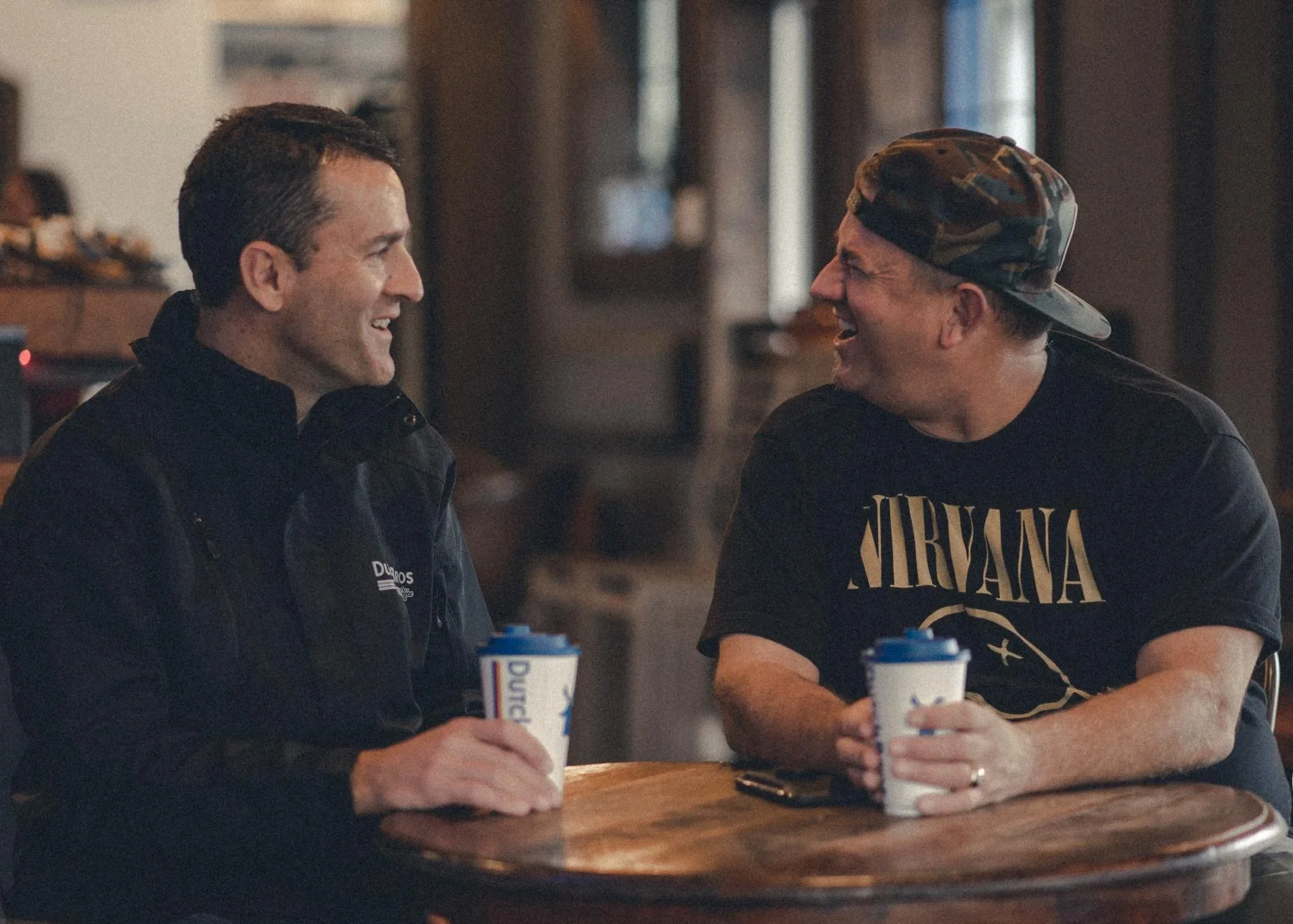 Two men laughing and enjoying coffee together; one in dark jacket, other wearing camouflage cap and Nirvana t-shirt.