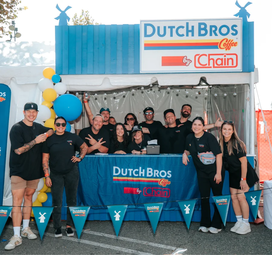Dutch Bros Coffee stand with staff in black shirts posing together in front of blue counter with colorful balloons and pennants.