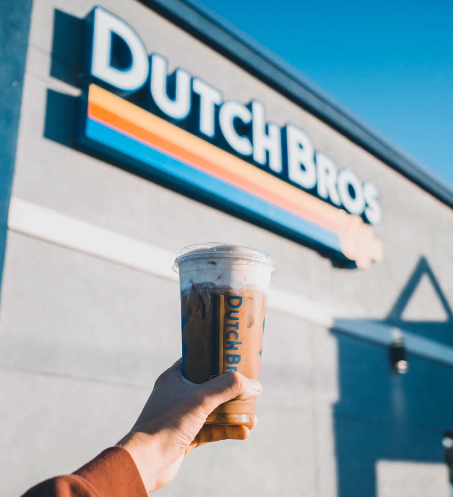 Hand holding an iced coffee drink in front of a Dutch Bros coffee shop with its distinctive blue and orange logo.