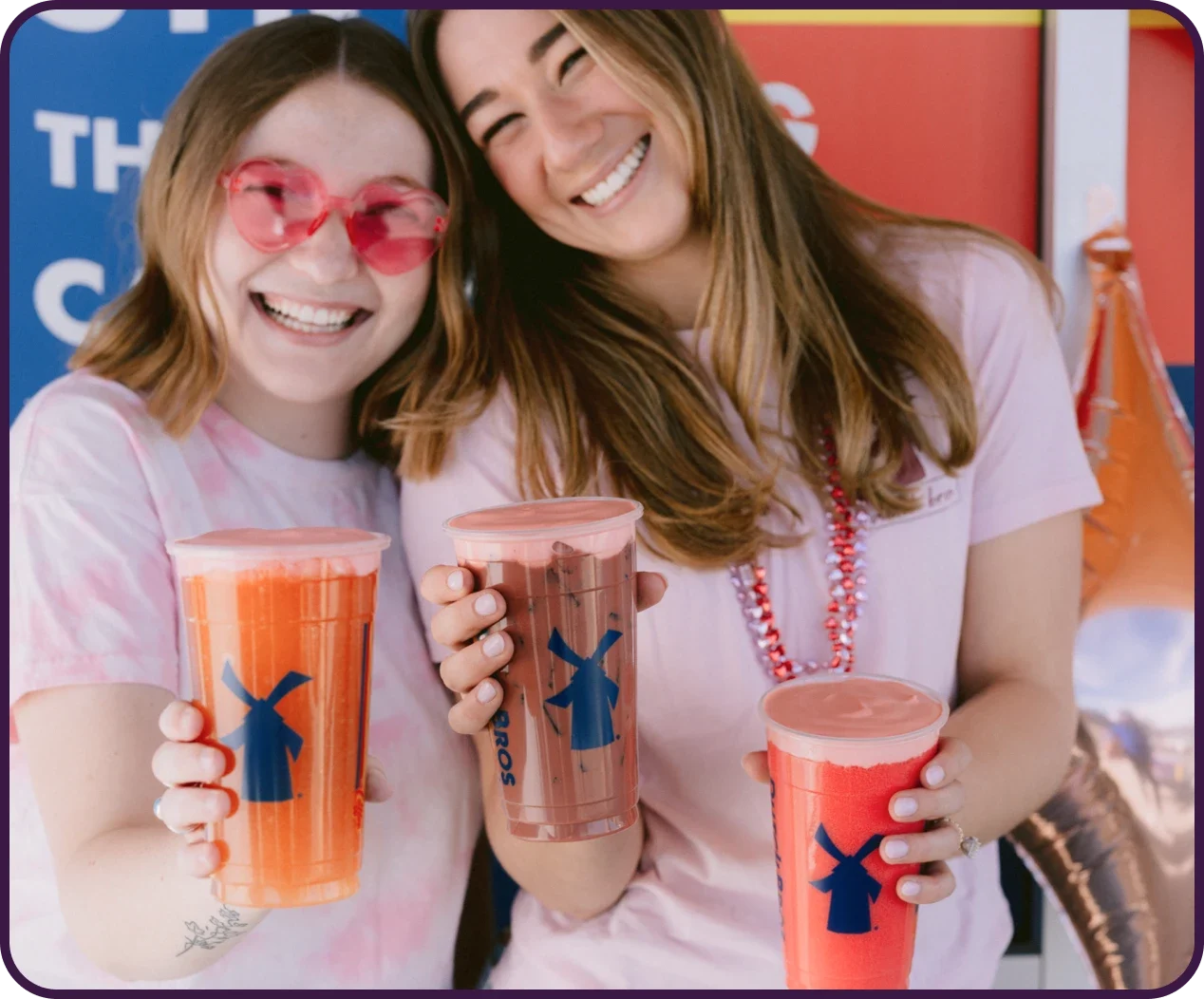 Two smiling friends in pink shirts holding colorful Dutch Bros drinks with windmill logos against a blue and red background.