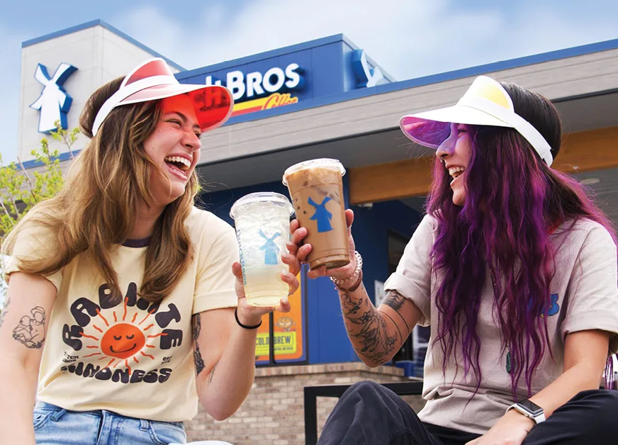 Two friends in sun visors toasting iced coffee drinks outside a Dutch Bros Coffee shop on a sunny day.
