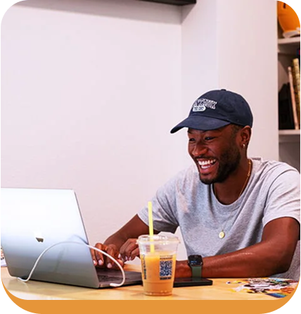 Person in blue cap smiling while working on laptop at wooden table with iced drink nearby