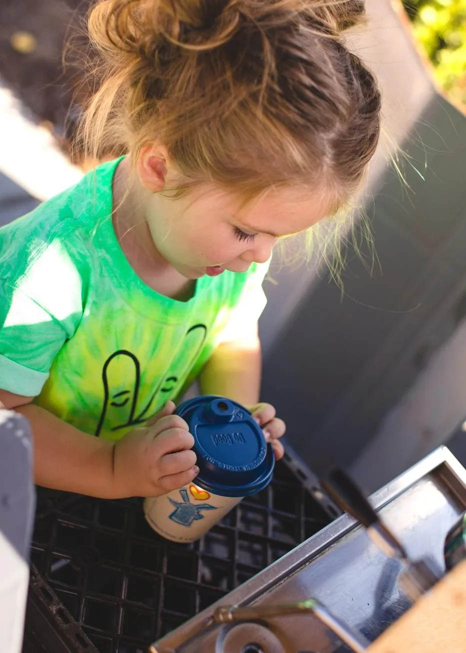 Child in bright green tie-dye shirt holding a paper cup with blue lid, looking down intently.