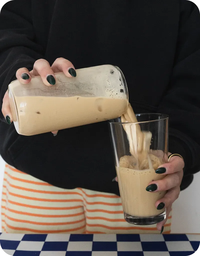 Person with dark green nails pouring creamy iced coffee from jar to glass, wearing black top and striped skirt.