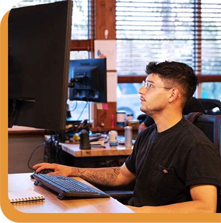Person with glasses working at computer desk with keyboard and monitor in office setting with wooden blinds.