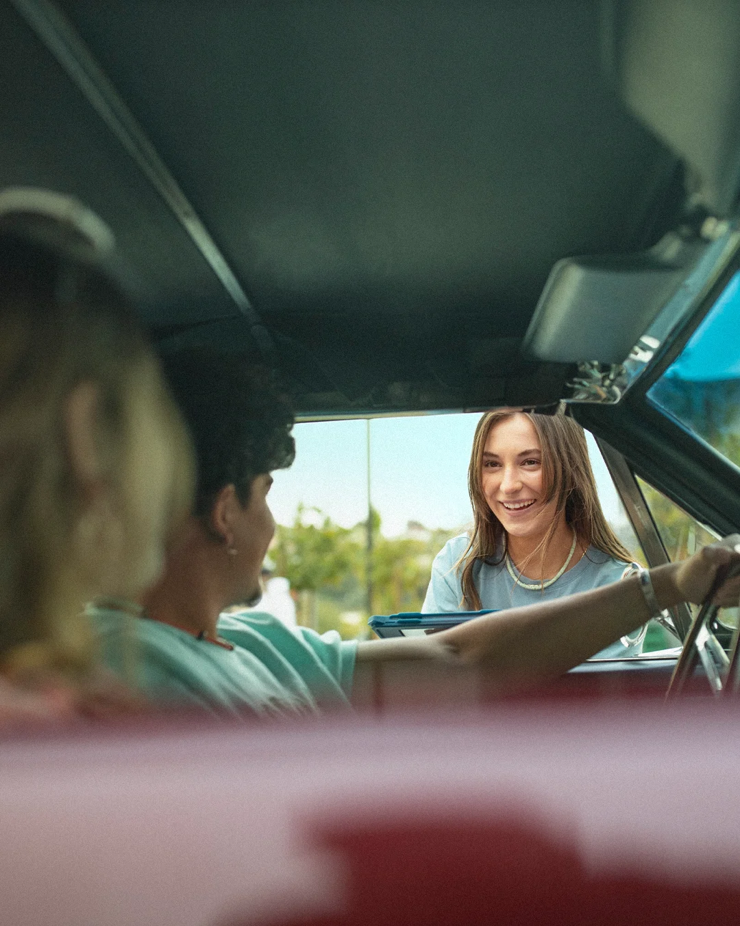 View from inside a car as someone in a light blue shirt smiles through the open window on a sunny day.