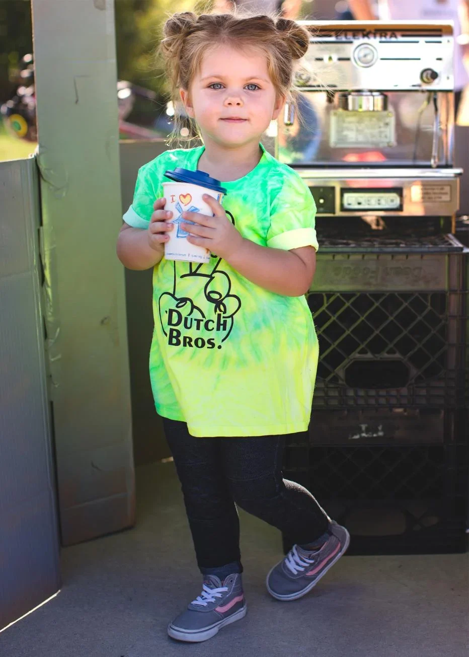 Young child in bright green Dutch Bros shirt holding a coffee cup, standing next to coffee equipment.