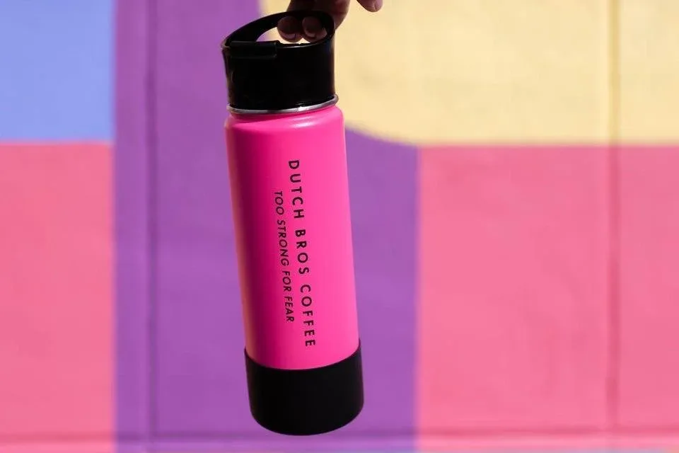 Hand holding a bright pink Dutch Bros Coffee insulated travel mug against a colorful pink and purple background.