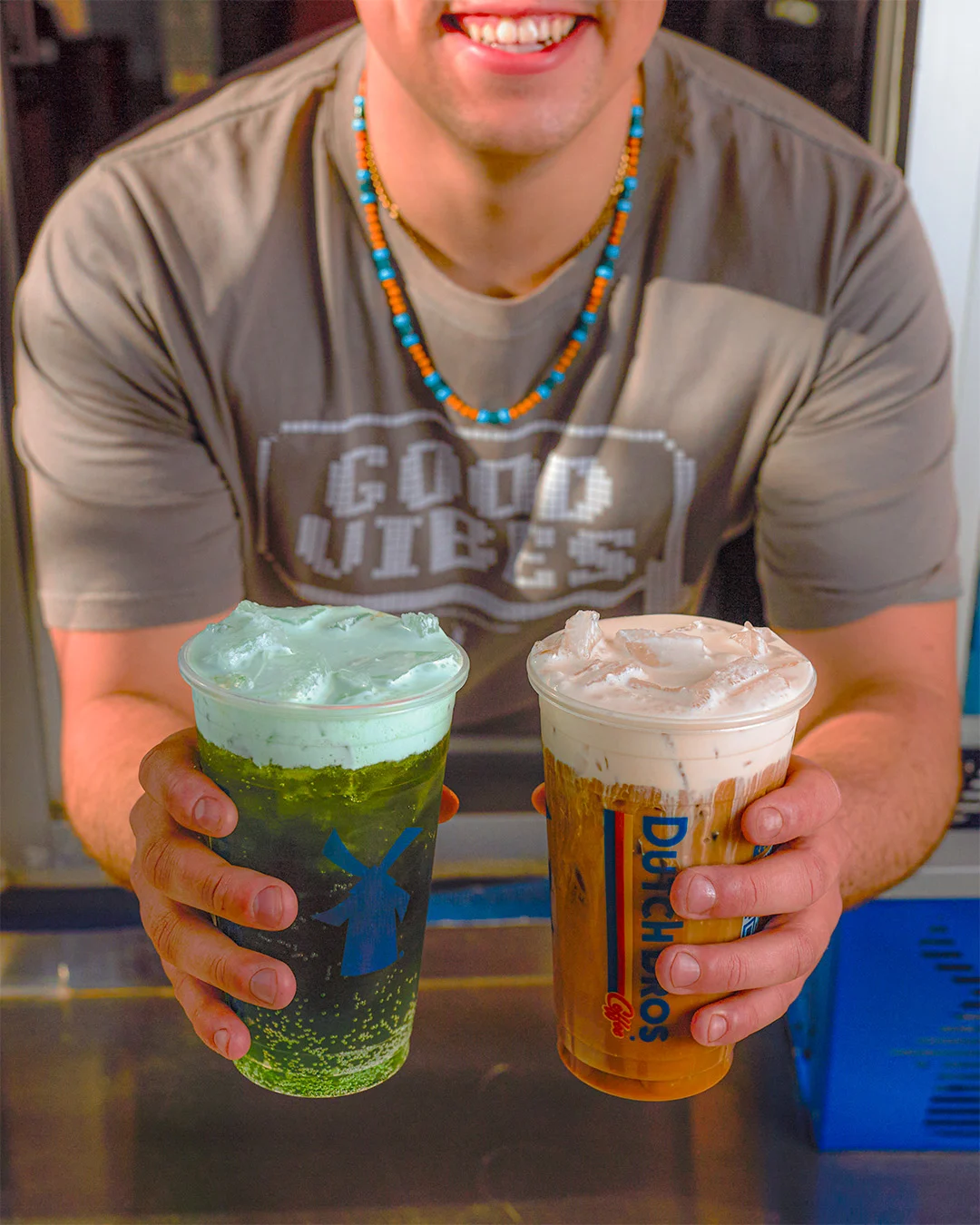 Person wearing beaded necklace holding two colorful bubble tea drinks - one green and one brown with creamy tops.