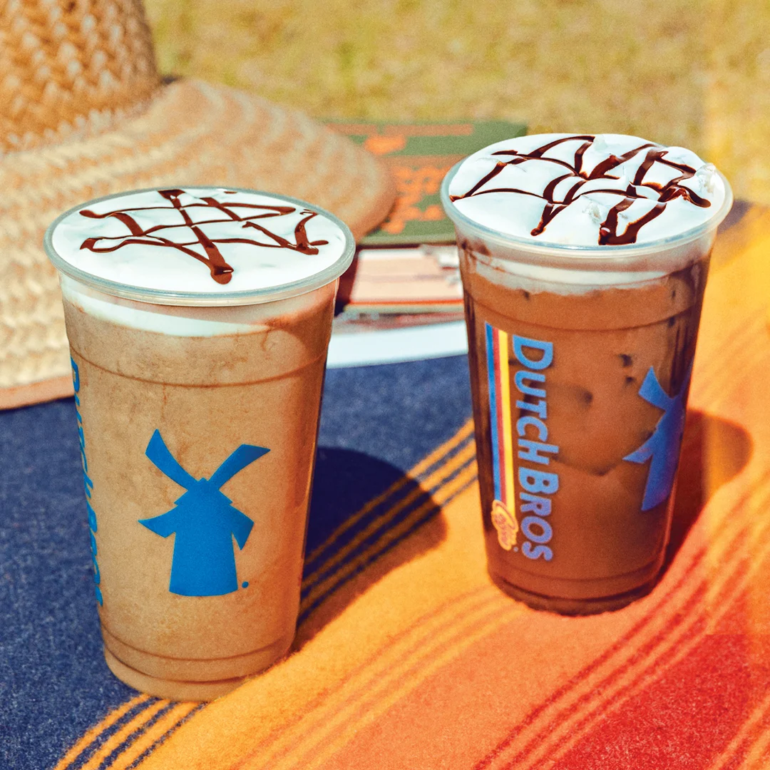 Two Dutch Bros iced coffee drinks with whipped cream and chocolate drizzle on a colorful table.