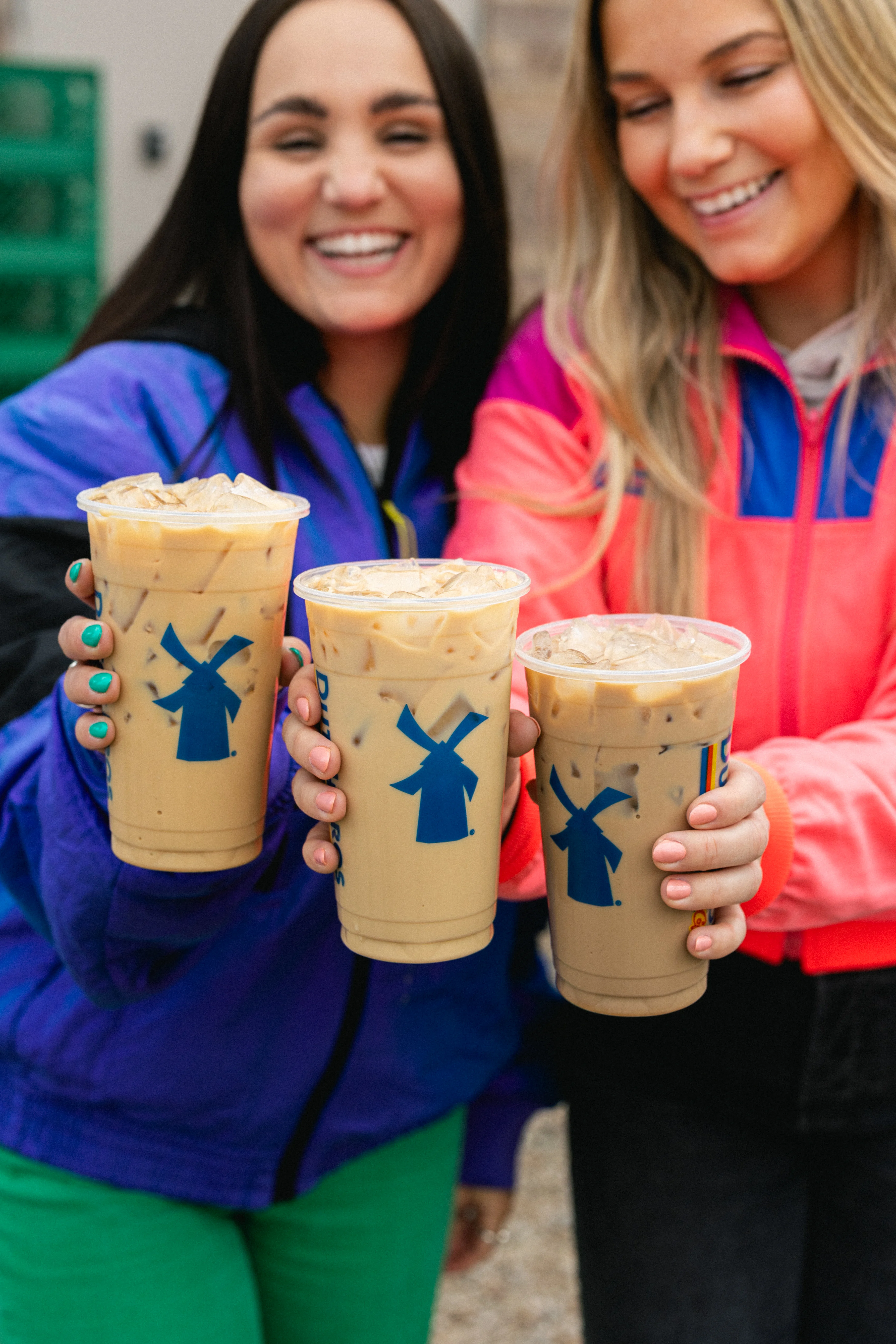 Two smiling friends in colorful jackets holding iced coffee cups with blue windmill logos.