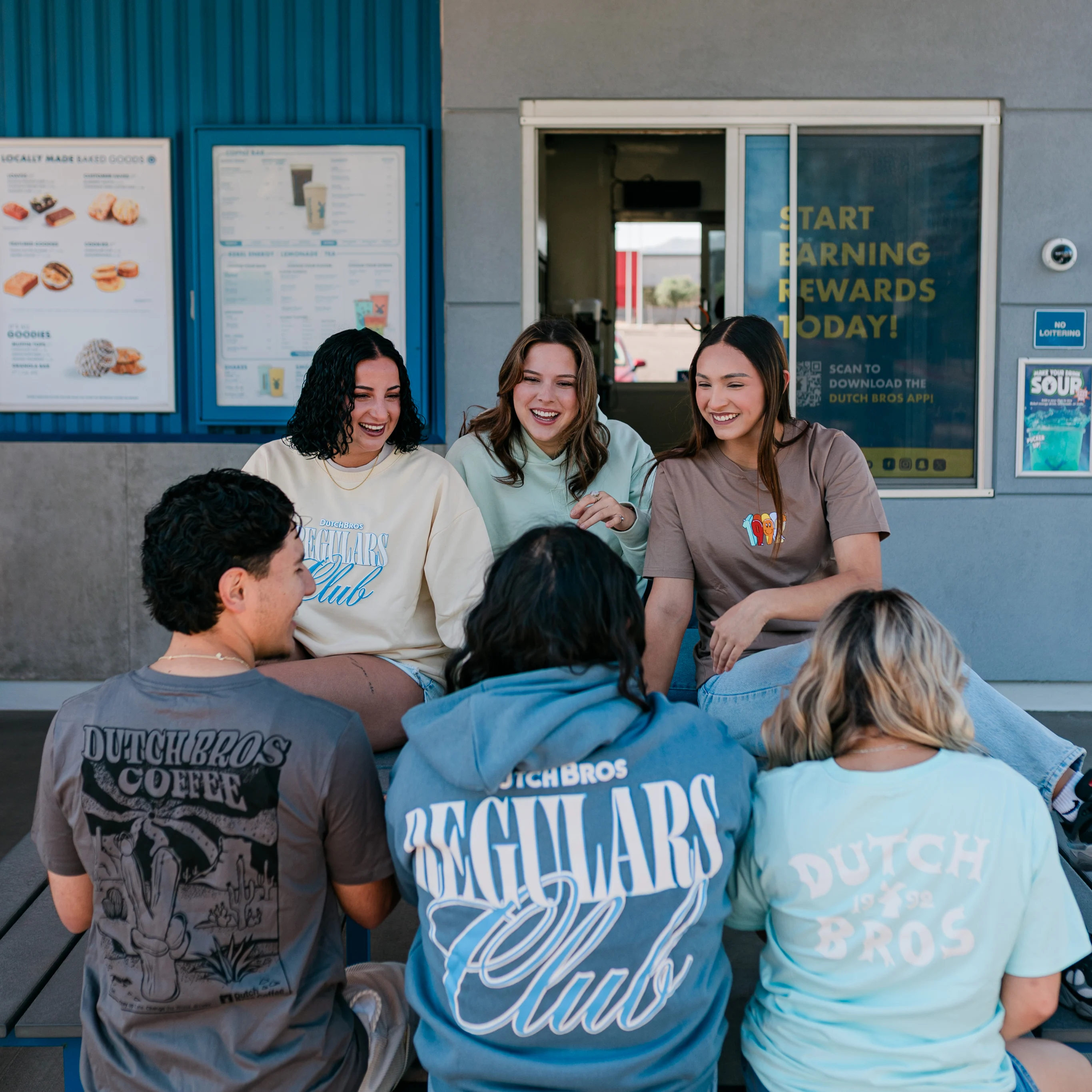 Friends laughing together outside Dutch Bros coffee shop, wearing branded merchandise with rewards sign visible.