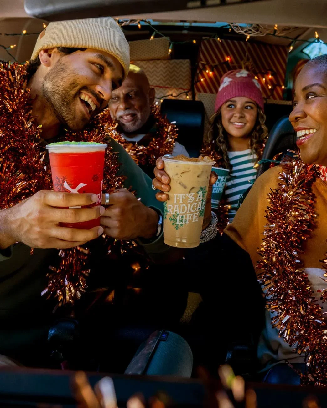 Friends celebrating in a car with festive tinsel garlands, holding holiday drinks, wearing winter hats under string lights.