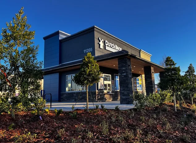 Dutch Bros coffee shop building with blue exterior, stone accents, and landscaped surroundings against clear blue sky.