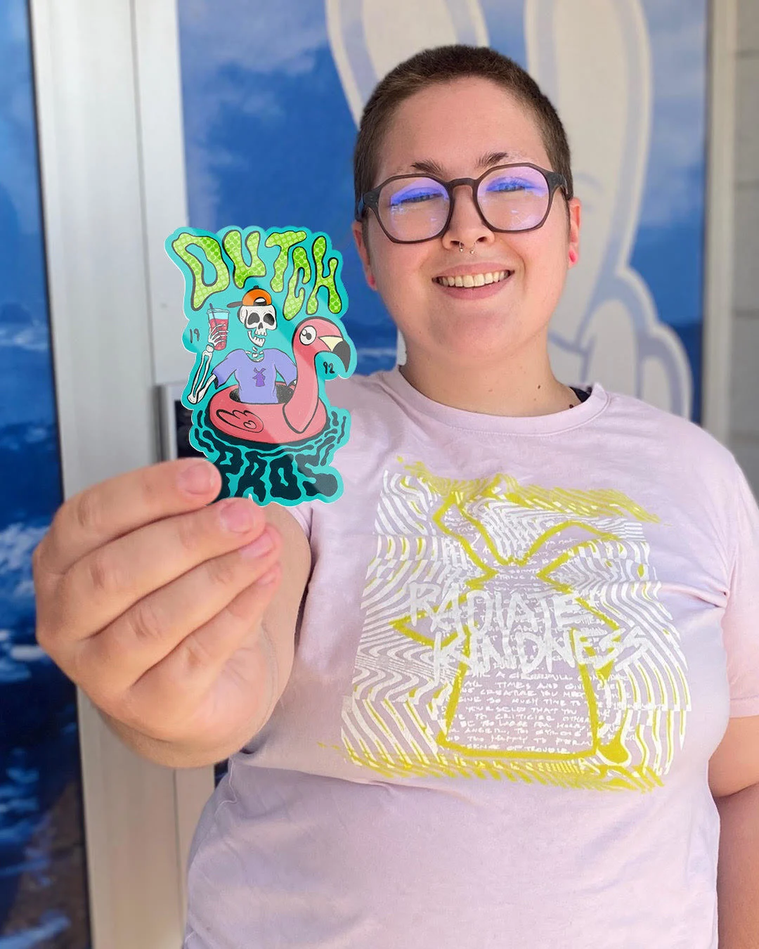 Person with glasses displaying a colorful "Dutch Bros" sticker featuring a skeleton on a flamingo float against blue background.