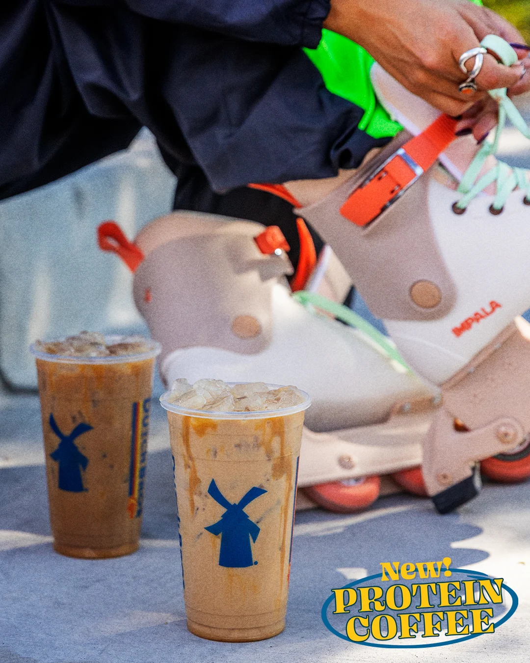 Two iced coffee cups with blue windmill logos beside colorful rollerskates, advertising "New Protein Coffee"