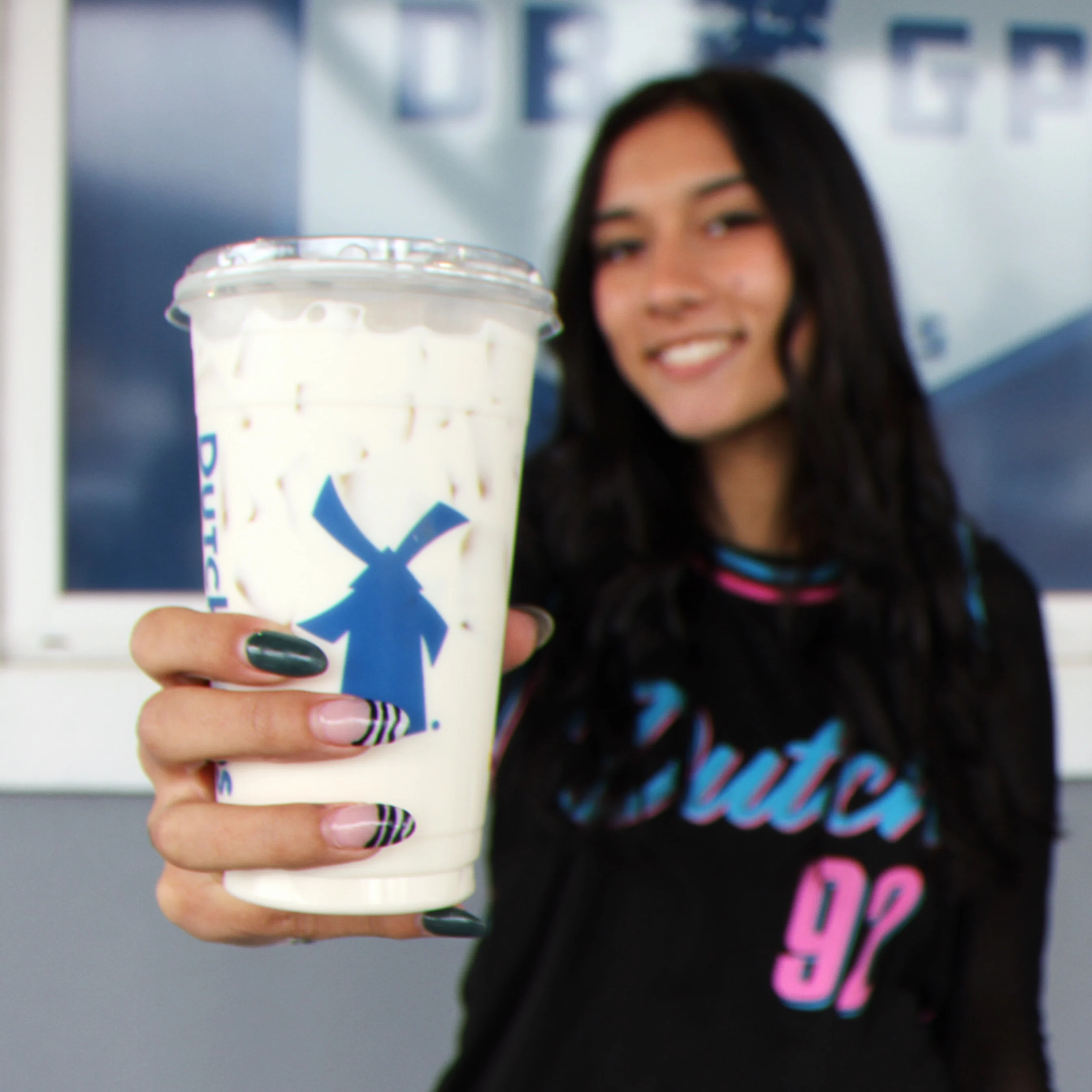 Person holding a Dutch Bros coffee cup with blue windmill logo, wearing colorful graphic t-shirt with styled nails.