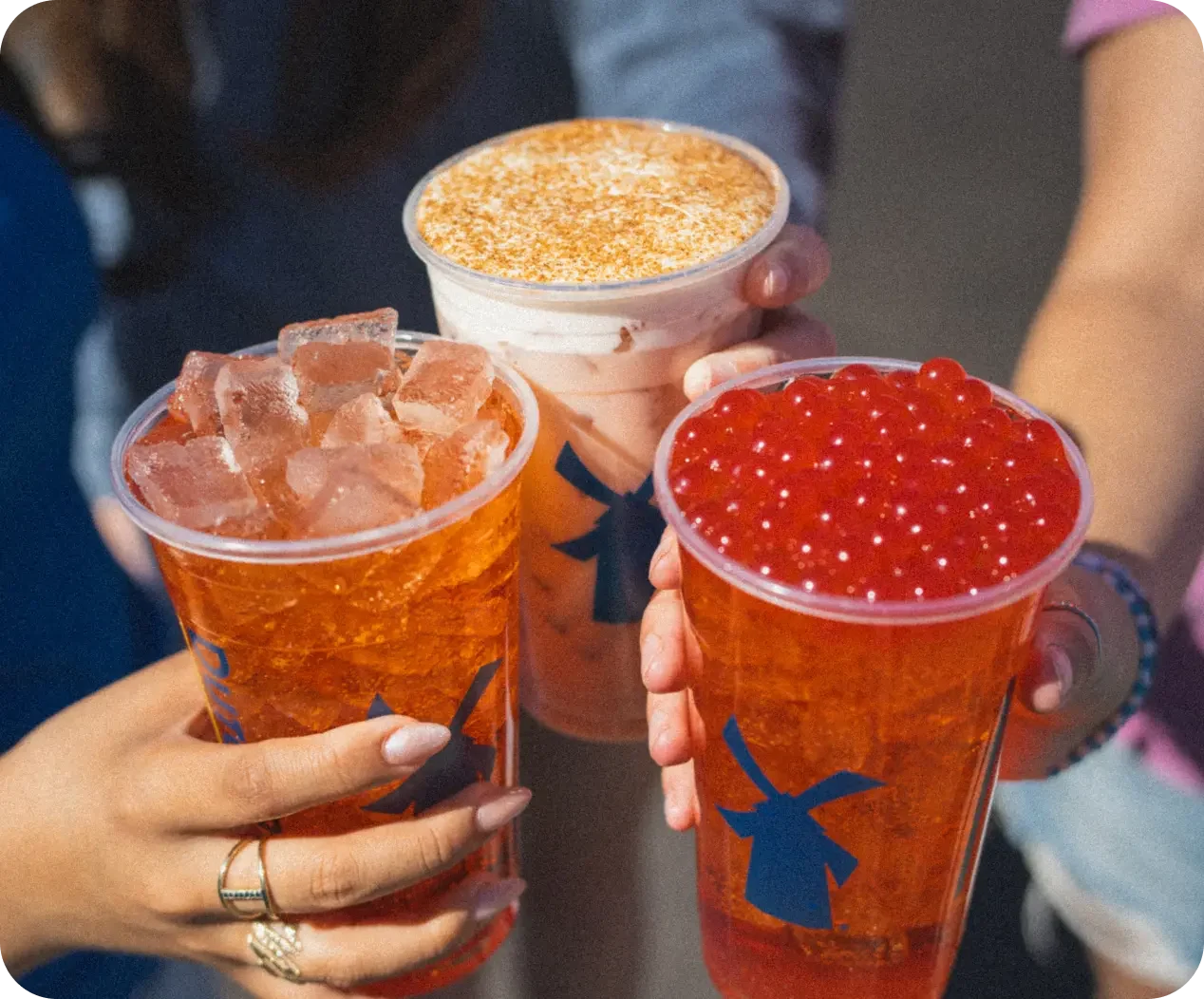 Three hands holding Dutch Bros drinks: an iced tea, a creamy topped beverage, and a red boba drink.