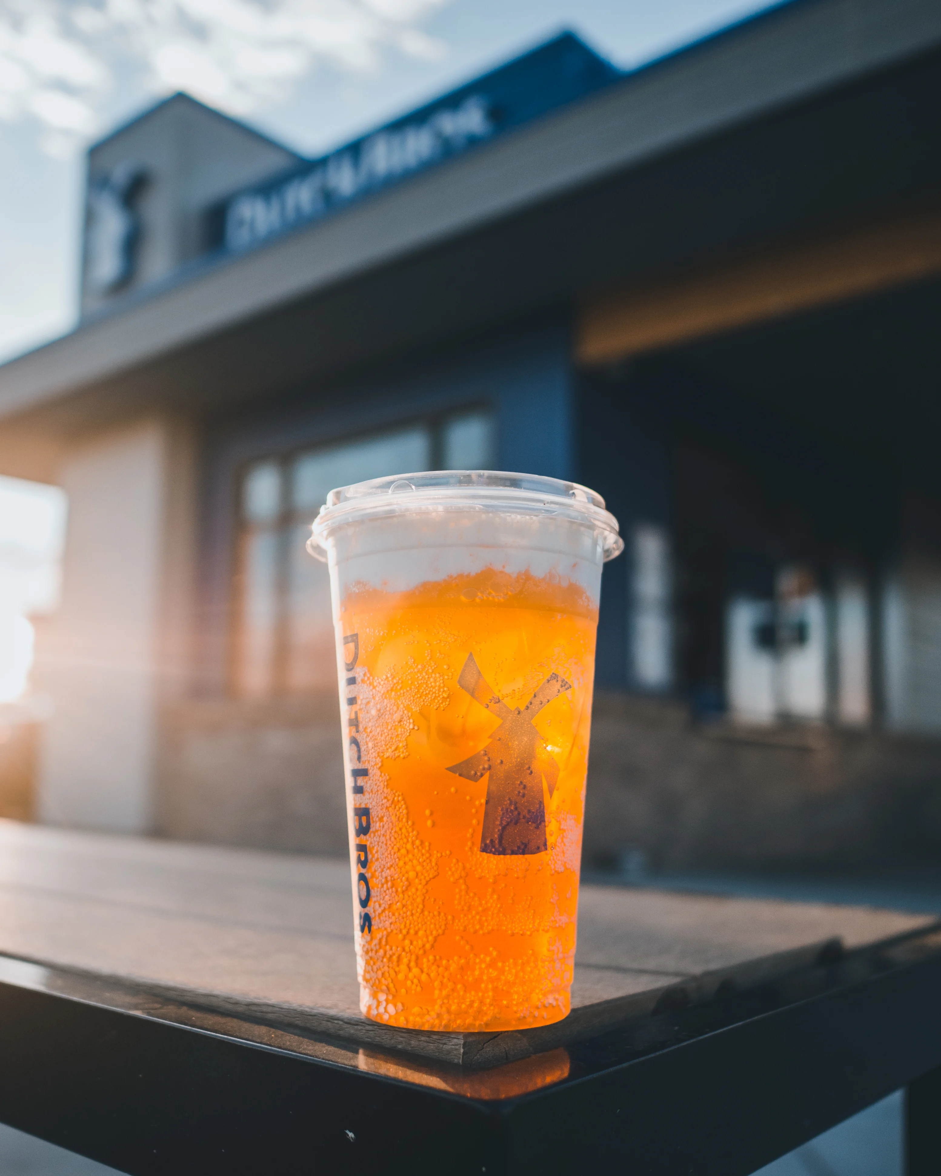 Orange Dutch Bros beverage in clear cup with windmill logo, storefront visible in background.