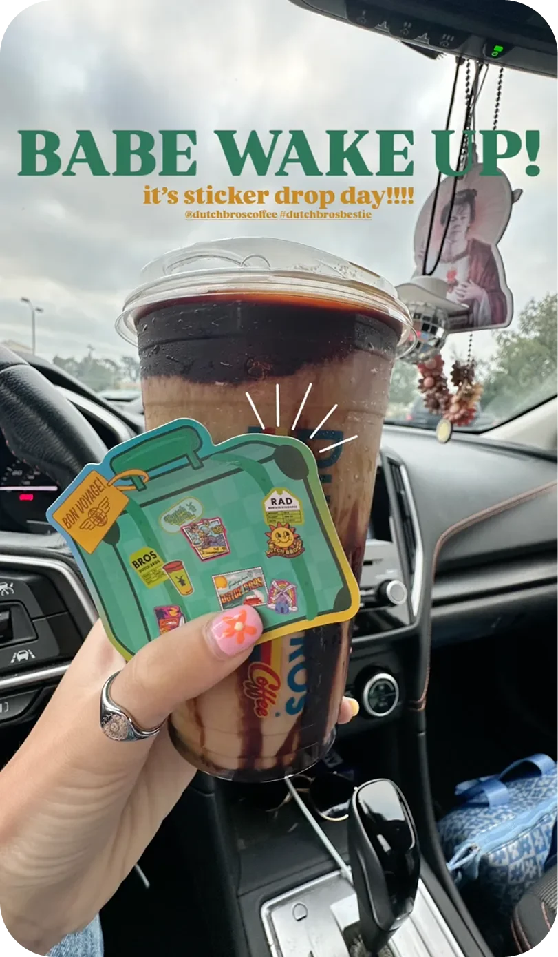 Hand holding a Dutch Bros iced coffee and a suitcase-shaped sticker inside a car, with text reading "Babe Wake Up! It's sticker drop day!"