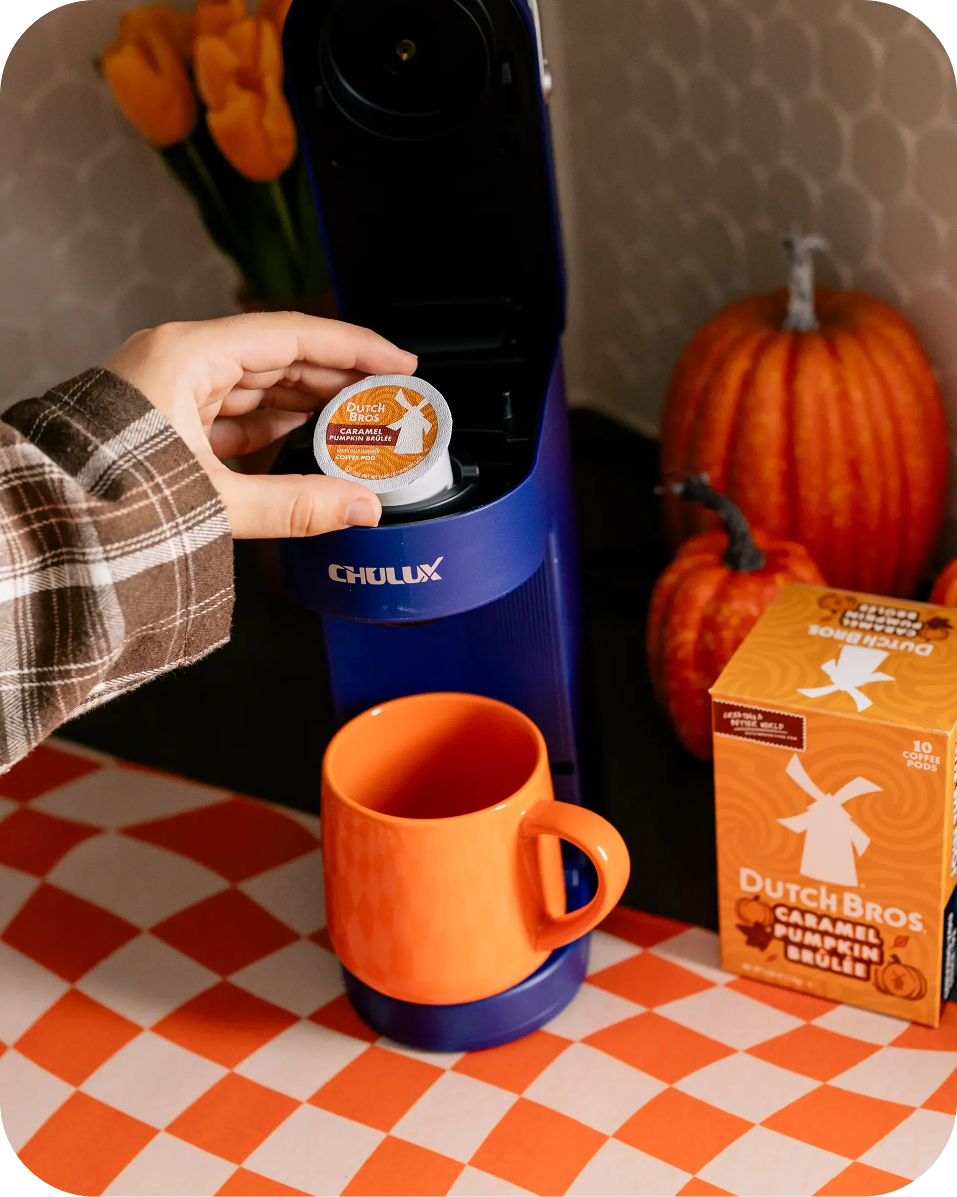 Hand inserting a Dutch Bros Caramel Pumpkin Brûlée coffee pod into a blue Chulux machine beside an orange mug and pumpkins.