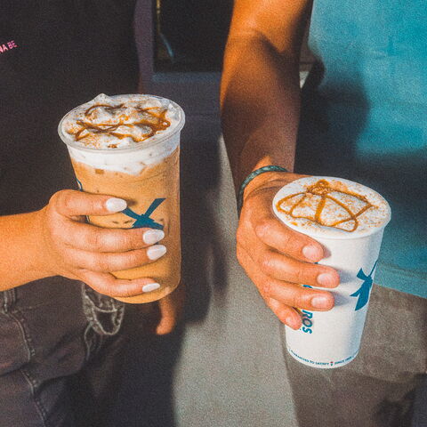 One persons hand holding an iced coffee with soft top and drizzle with no lid and another hand holding a warm drink with no lid