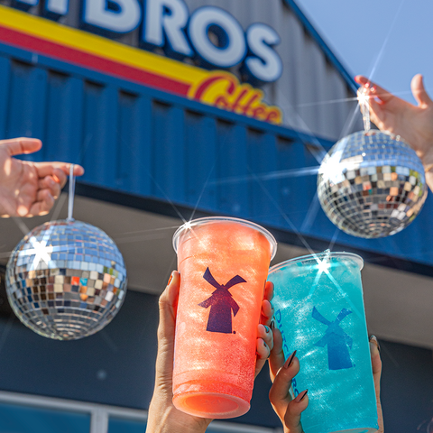 two hands holding up two dutch bros drinks with two discos shining in the sun and in front of a dutch bros shop