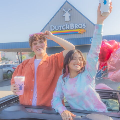 Two individuals standing out of a car sunroof with a Dutch Bros in hand